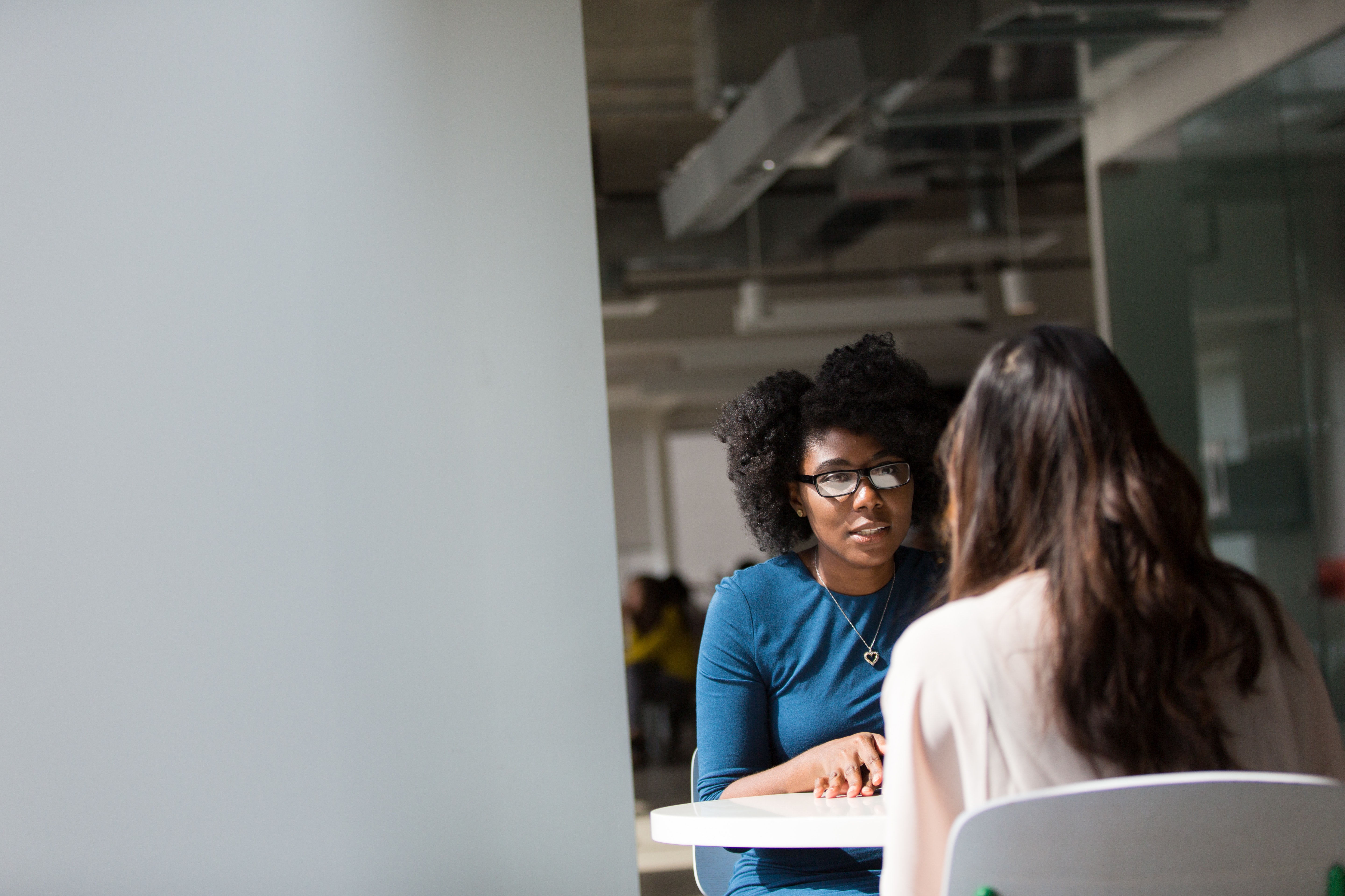 Two women sit at a white table in an office building, facing each other and talking